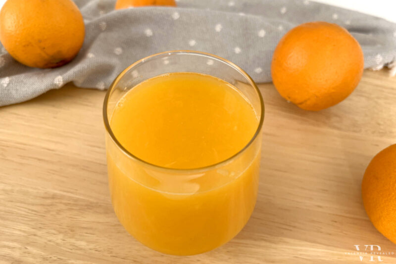 A glass of freshly squeezed orange juice on a table with a cloth and three oranges